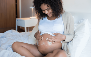 gravid kvinne sitter i en seng og smører magen sin med krem
