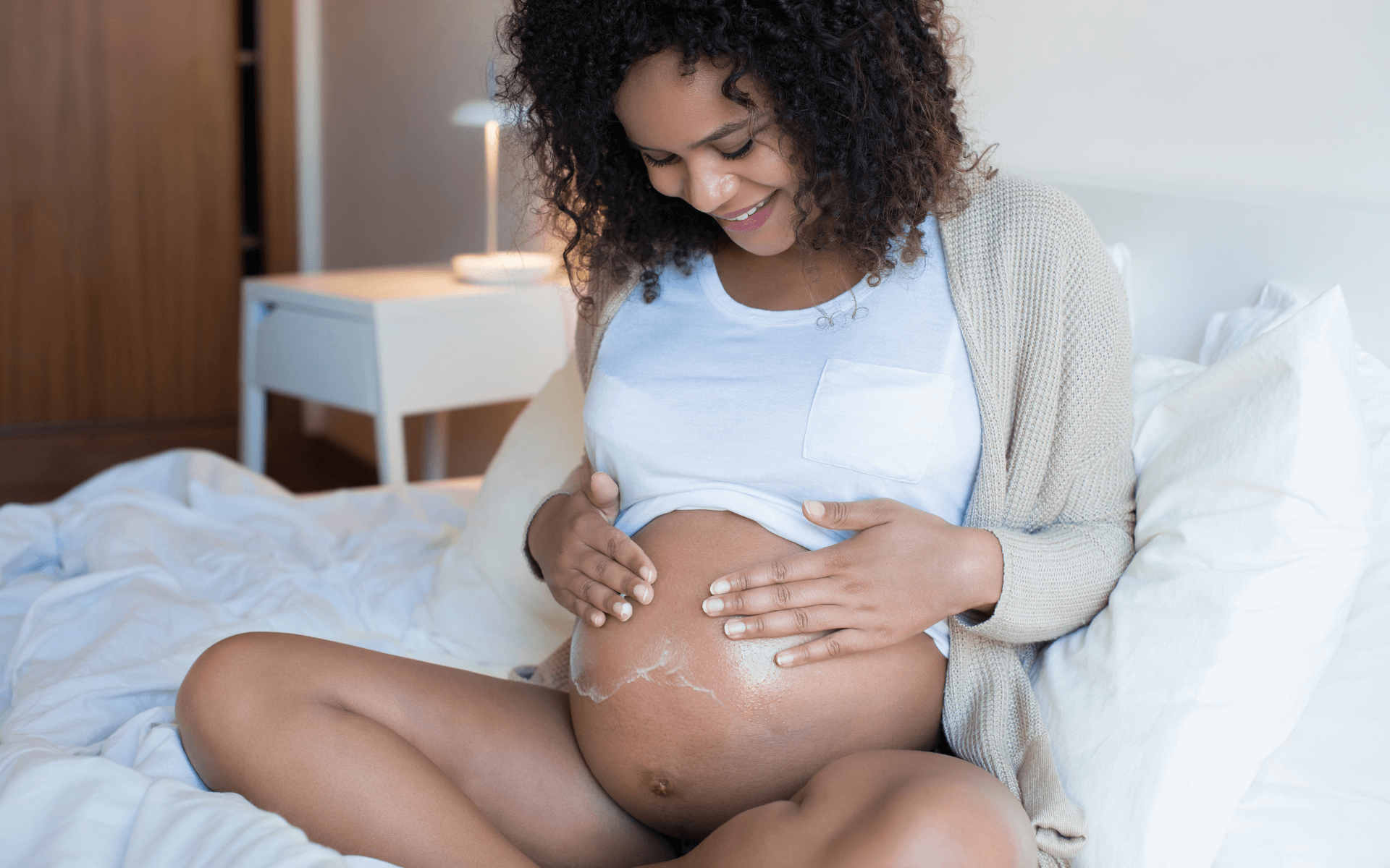 gravid kvinne sitter i en seng og smører magen sin med krem