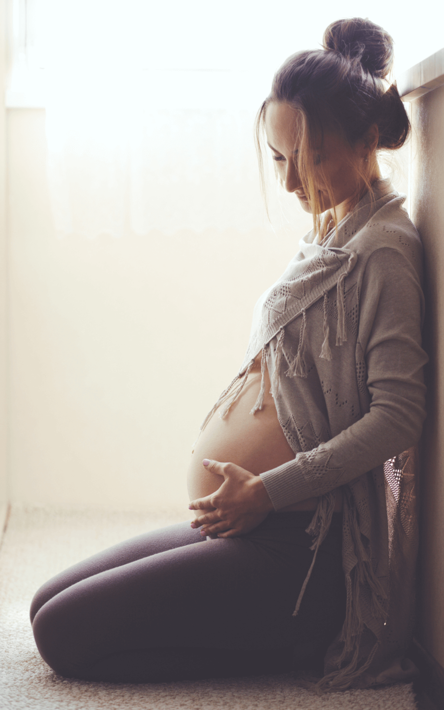 Gravid kvinne med hendene på magen sitter på gulvet og smiler og ser ned på magen sin